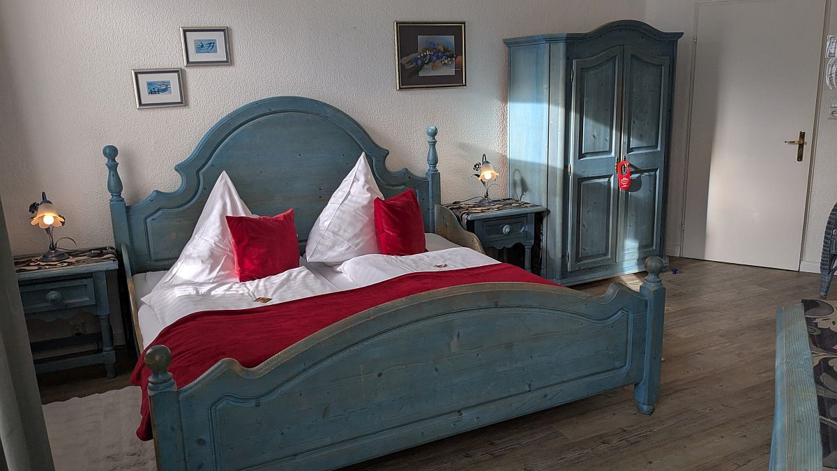 Interior view of a double room at Hotel Jägerhof. In the center of the room is a double bed with red pillows and a red cozy blanket. On both the right and left sides of the bed, there is a bedside table with a bedside lamp.