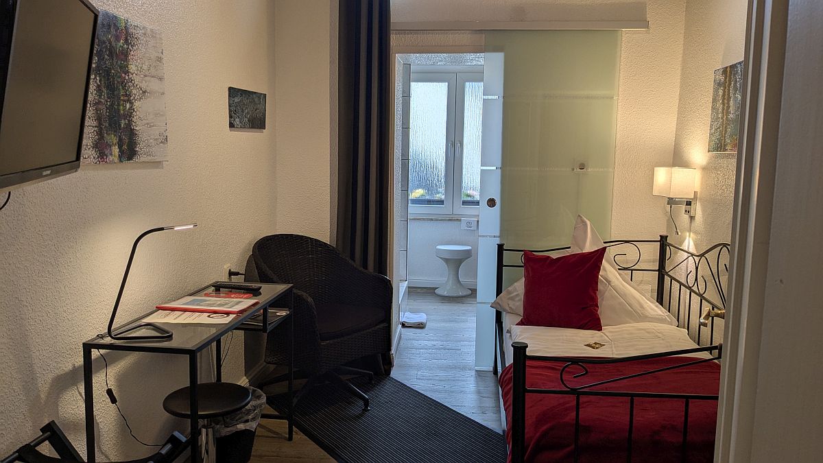 Interior view of a single room at the Hotel Jägerhof in Mayen. The room has a single bed, a desk and a television. The room has its own bathroom with a shower and cosmetic mirror. Rooms are cleaned daily.
